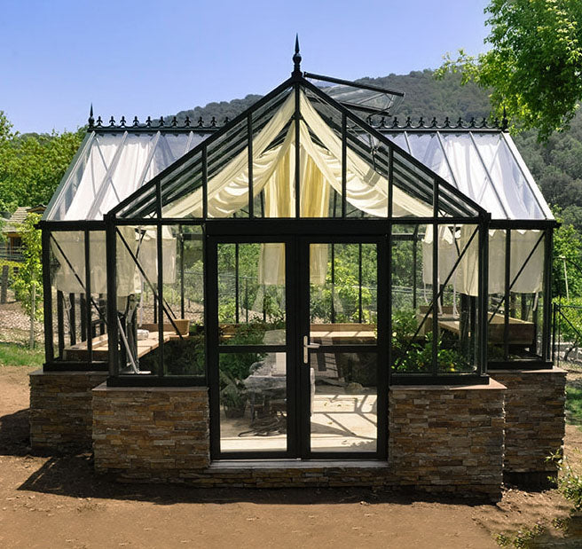 Elegant Exaco Janssens Royal Antique Orangerie Greenhouse with a stone base, surrounded by lush greenery and hills in the background, showcasing its classic design and spacious interior