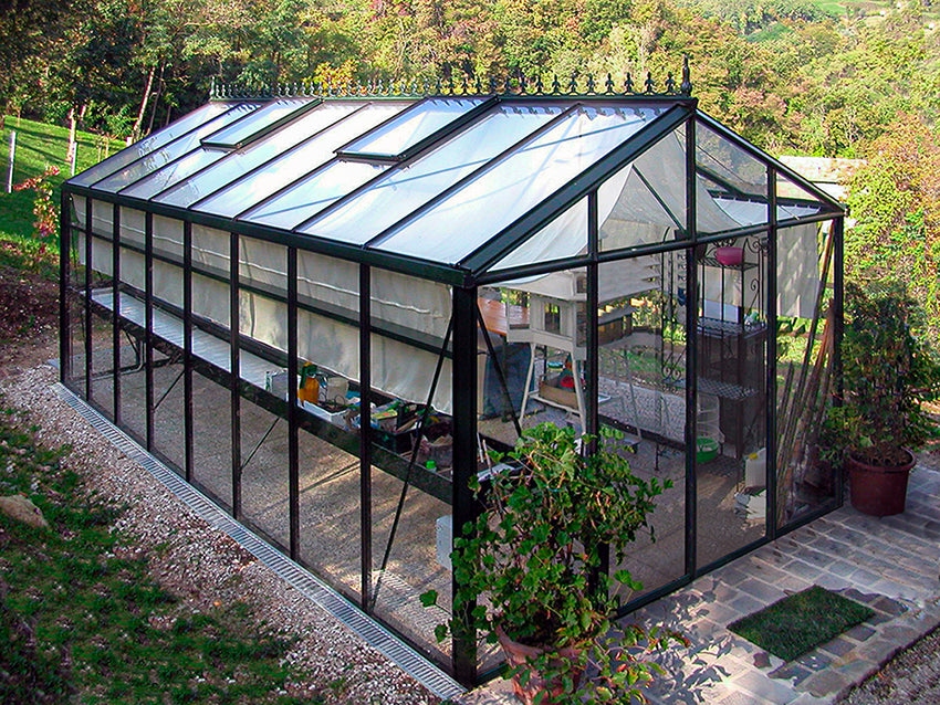 Exaco Royal Victorian VI 36 Greenhouse with sliding door in use, featuring spacious interior, durable glass panels, and elegant design, set in a lush garden with plants and gardening equipment inside. Perfect for year-round gardening.
