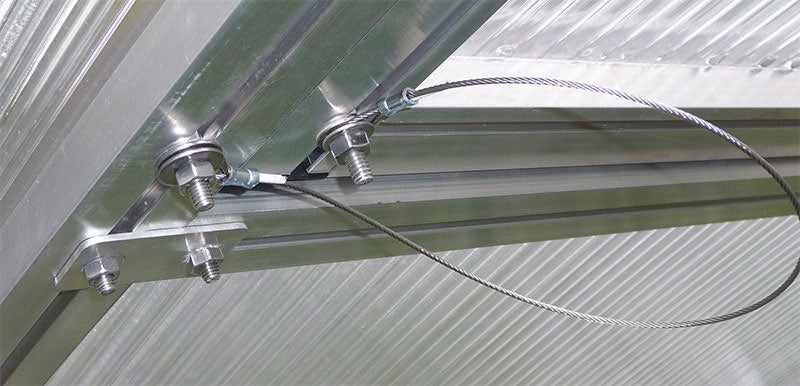 A close-up of a metal cable wind restraint system securing a greenhouse roof window.