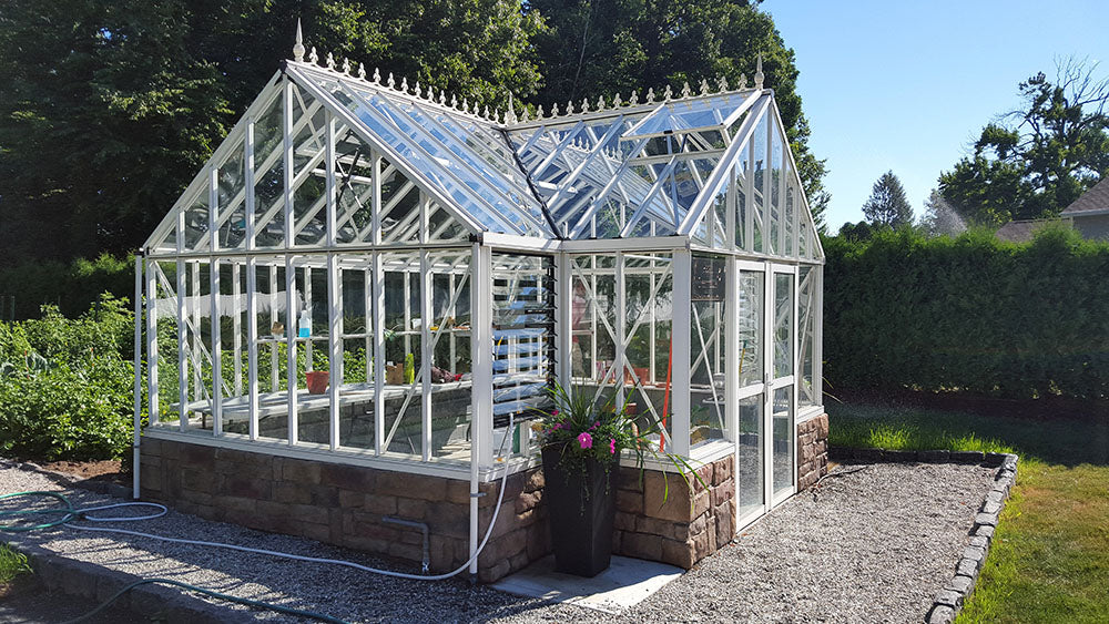 Classic white Exaco Janssens Royal Antique Orangerie Greenhouse set on a stone foundation next to a garden, illustrating its Victorian style and clear glass panels