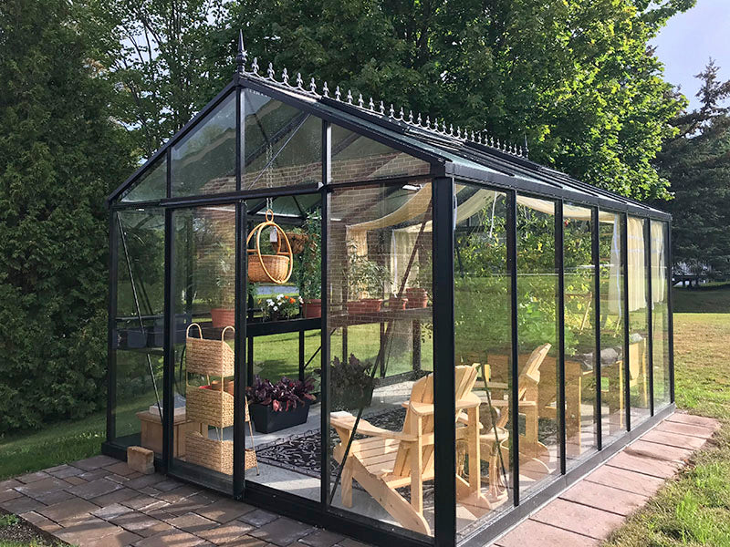 Black Royal Victorian greenhouse with glass panels and decorative roof cresting. Situated on a brick foundation, with wooden chairs and potted plants inside.