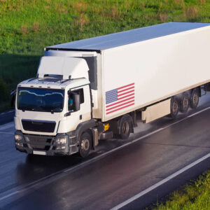 White semi-truck with an American flag on its trailer driving on a wet highway.