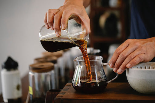 Hands pour coffee from a measuring cup into a pitcher.