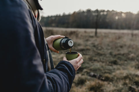 Person pouring a cup of coffee from a thermos while standing in a field