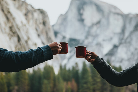 Two people raising coffee mugs together outside in the mountains.