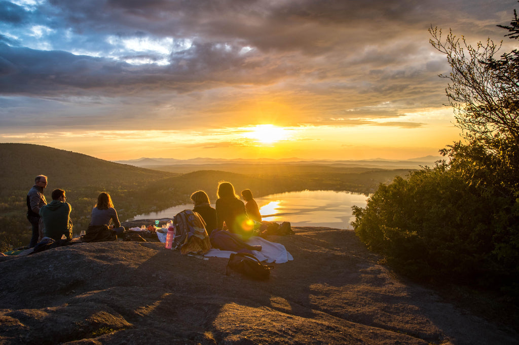 A large group watching the sun setting over a lake from a mountain ledge.