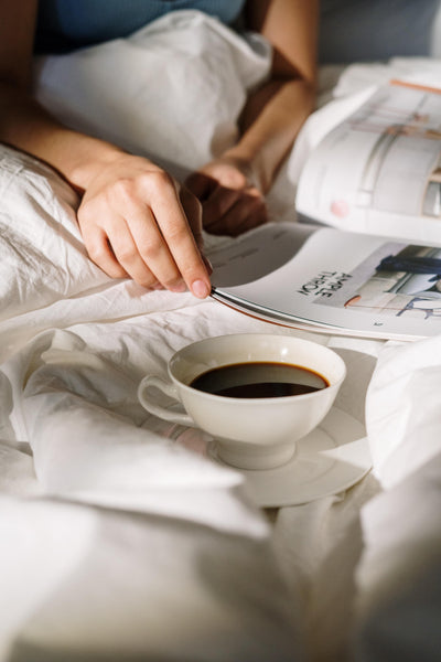 Hands flipping through magazine in bed with fresh coffee.