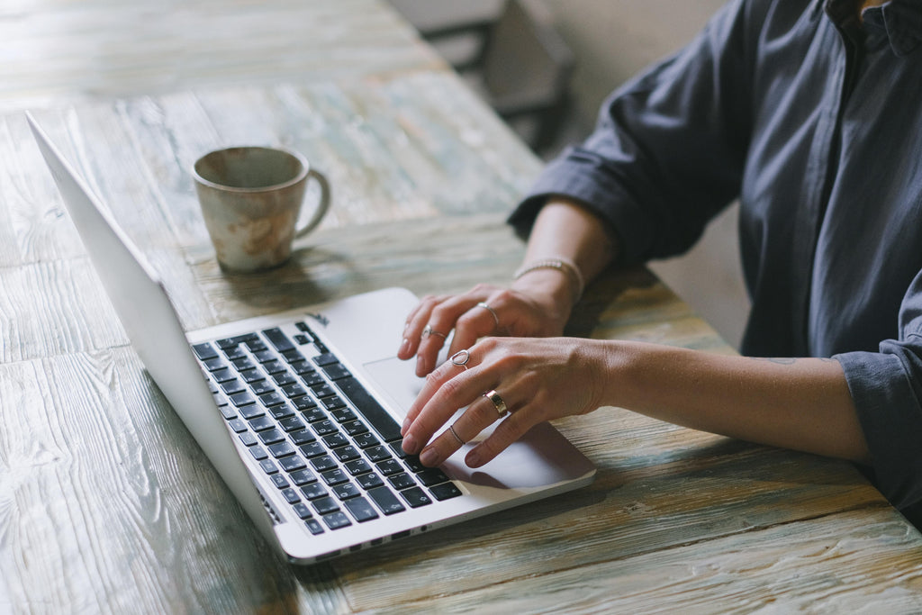 Hands typing at a laptop with a coffee cup on the table next to it. 