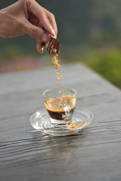 Hand pouring granules of brown sugar into a small espresso cup outdoors. 