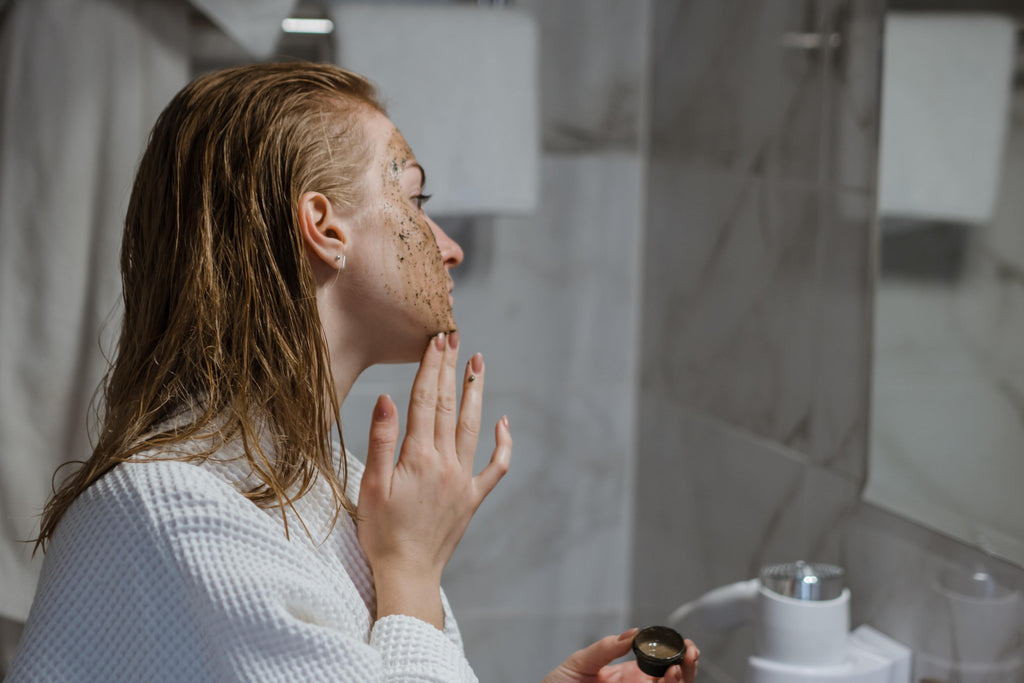 Woman scrubbing her face with a coffee scrub in front of mirror.