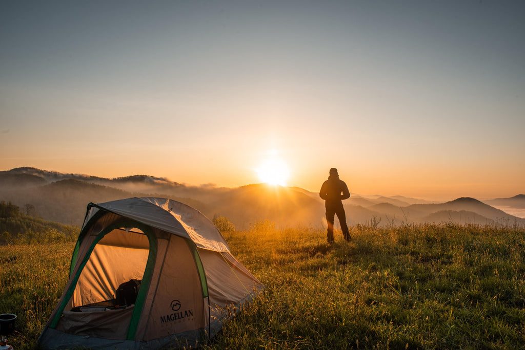 A tent is set up on top of a mountain, and a person in the distance watches the sunrise. 