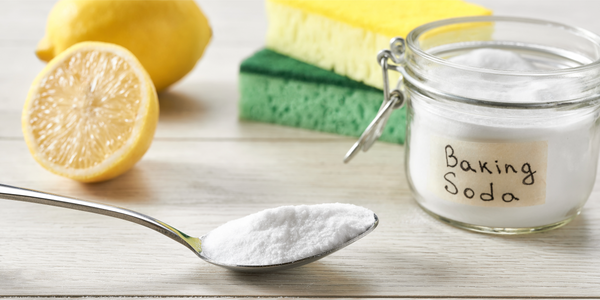picture of lemons, a green and yellow sponge, with a jar of baking soda and a spoonful of baking soda laying on counter.