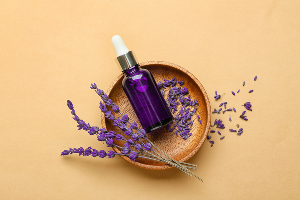 Image of a purple bottle laying in a wooden dish surrounded by lavender buds on a yellow background.