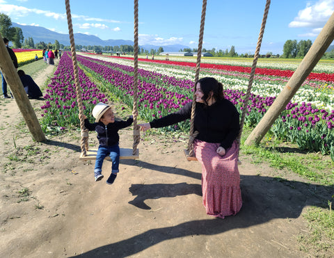 In a field of tulips a woman sits on a swing smiling at her son beside her who is looking at her on a swing also smiling 