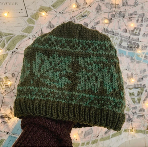 Two color hat held in one hand over a map, Gianna's first colorwork knitting project in forest and light green worsted weight yarn creating a walnut leaf pattern
