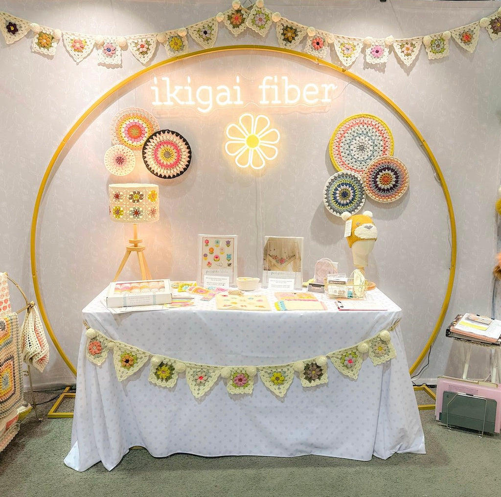 photo of ikigai fiber 2023 booth at h+h americas trade show. crochet bunting hangs above a neon logo.