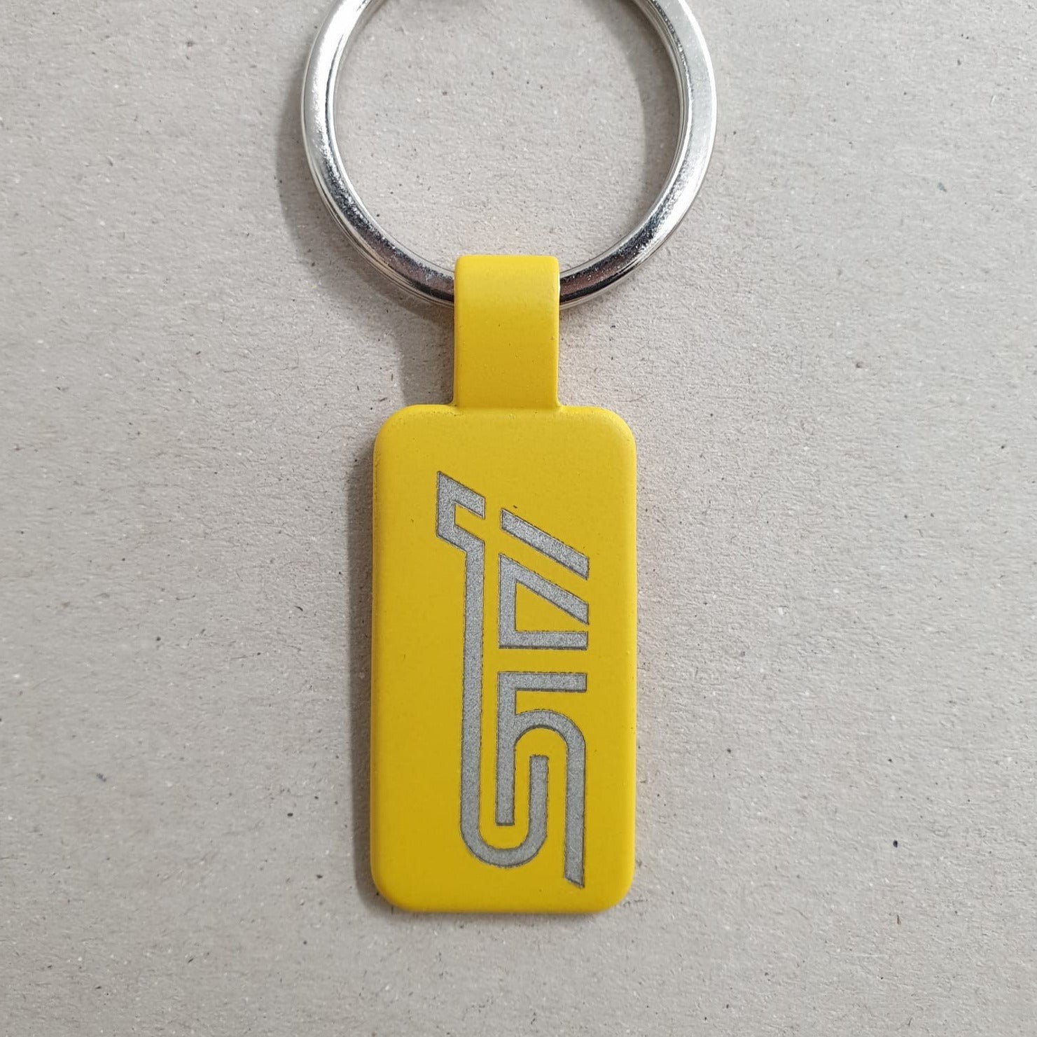 Llavero de acero, en color amarillo, con forma rectangular y anilla plateada, y grabado con el logotipo de Subaru STI , sobre fondo marrón texturizado.