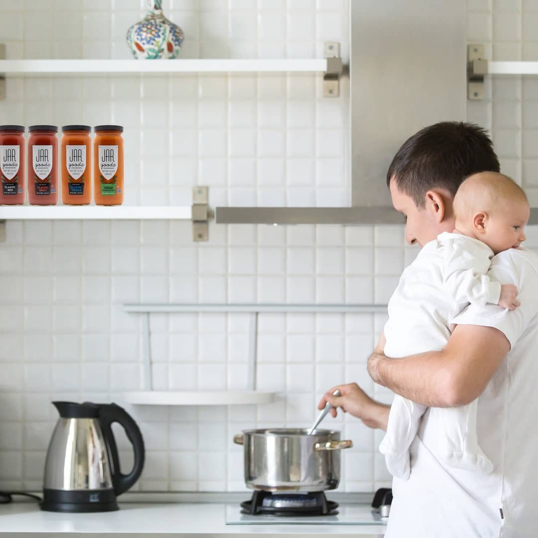 Parent making dinner with Jar Goods while holding his newborn baby.