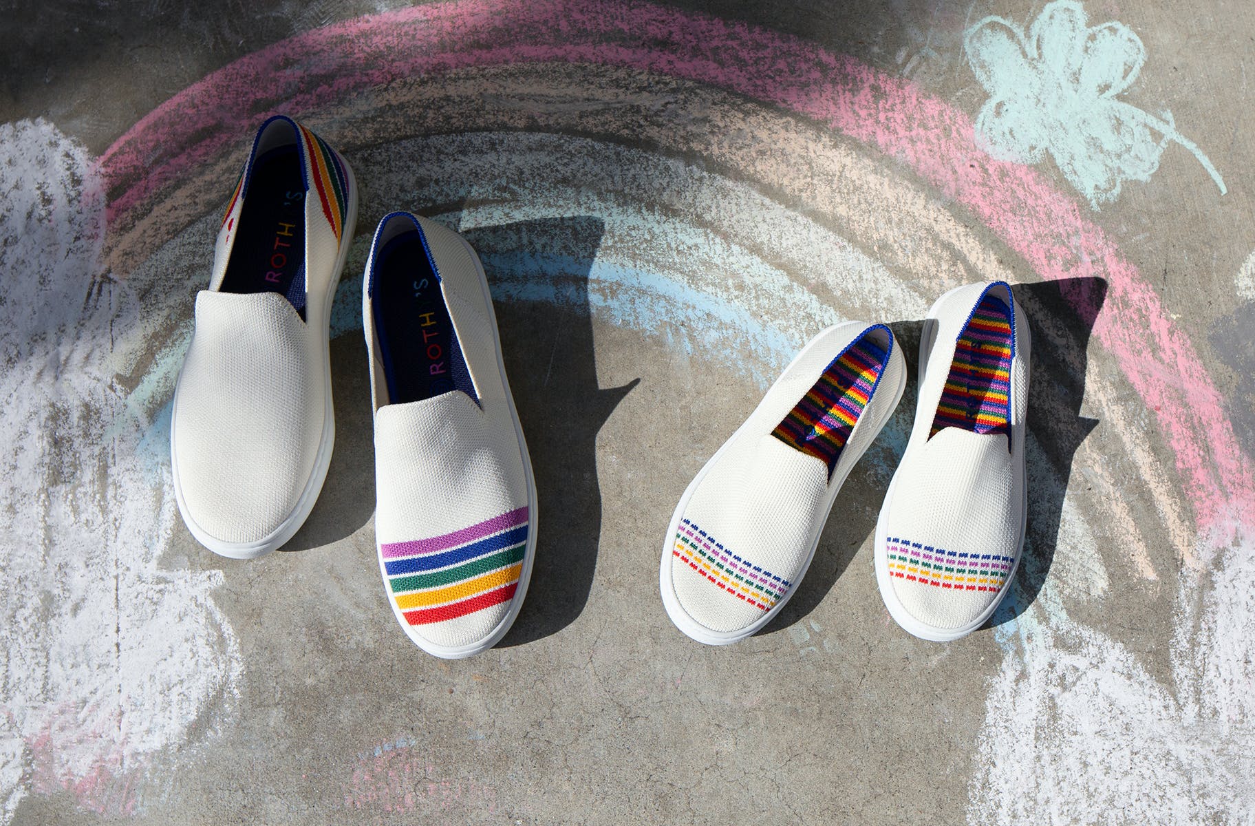 An adult's sneaker in white with multicolored stripes, and The Kids Sneaker in White Rainbow, shown on a sidewalk. 