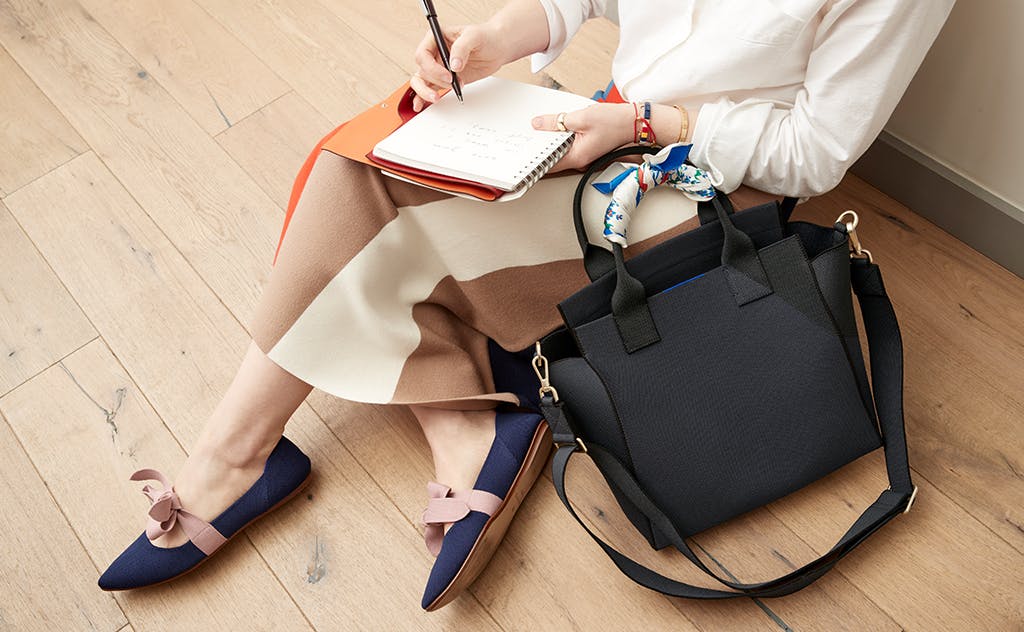 Art Director Gretchen sitting on a wooden floor while sketching in her notebook, with The Handbag in Midnight Navy next to her. 