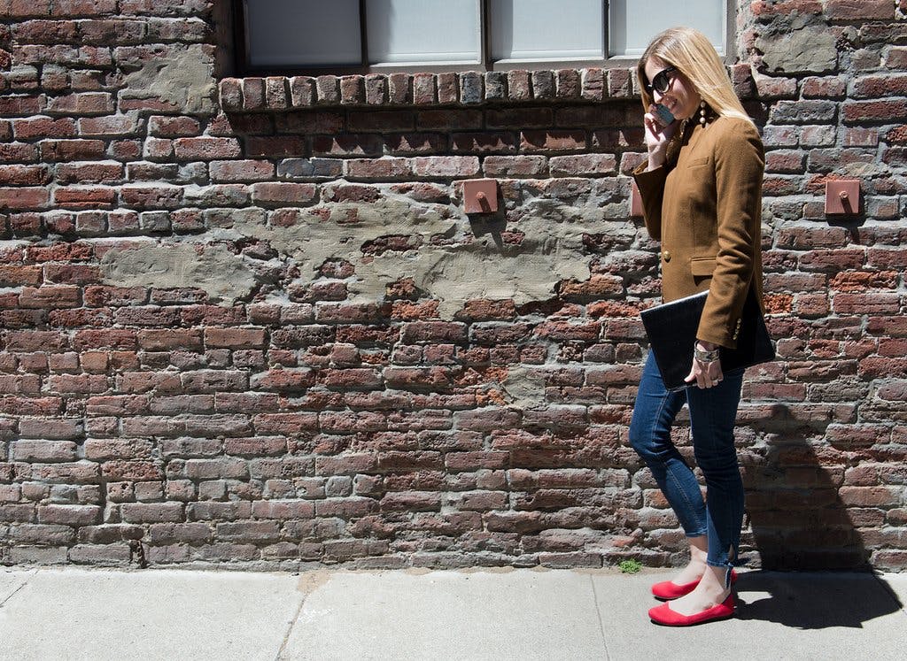 Woman shown in side profile wearing a pair of red Rothy's flats, standing in front of a red brick wall. 