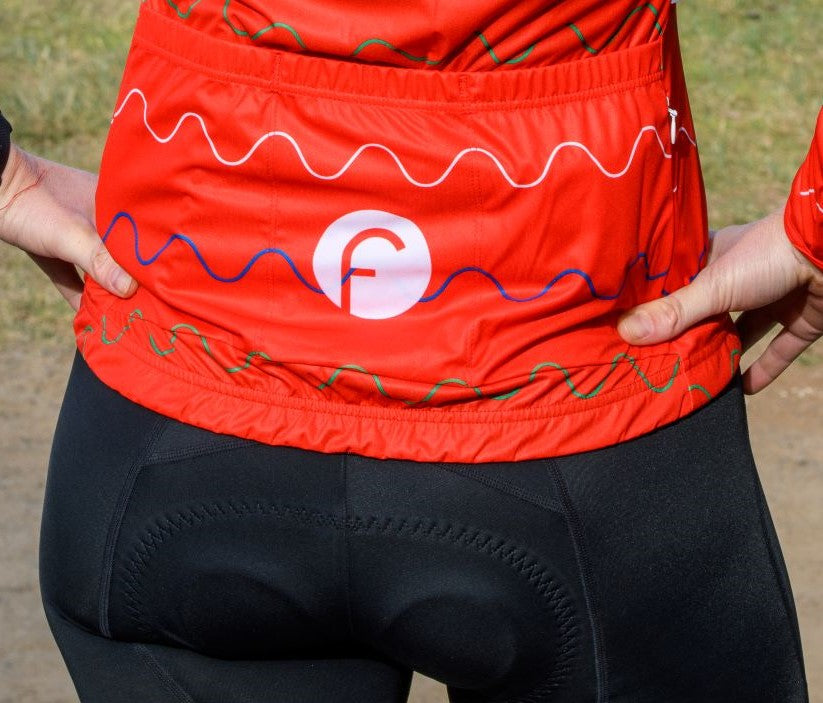 Close up view of women's cycling light packable jacket.  Shows light windproof and waterproof fabric with 3 large rear pockets to put your essentials including a waterproof zipper pocket on the side.  Fits easily over a long sleeve jersey or underlayer and packs down into a rear jersey pocket. Bright red colour for extra visibility.