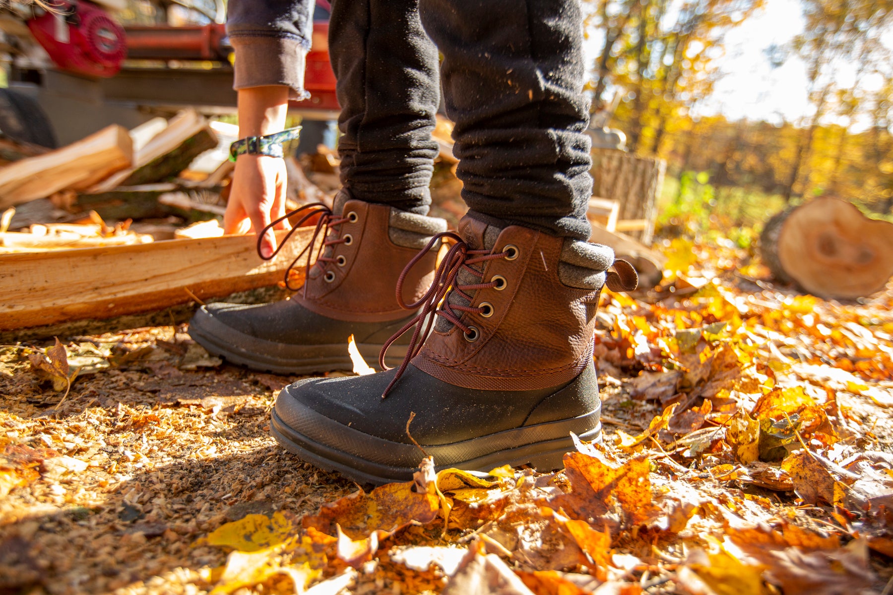 A person in a pair of boots reaching down for a piece of wood with Autumn leaves on the ground.