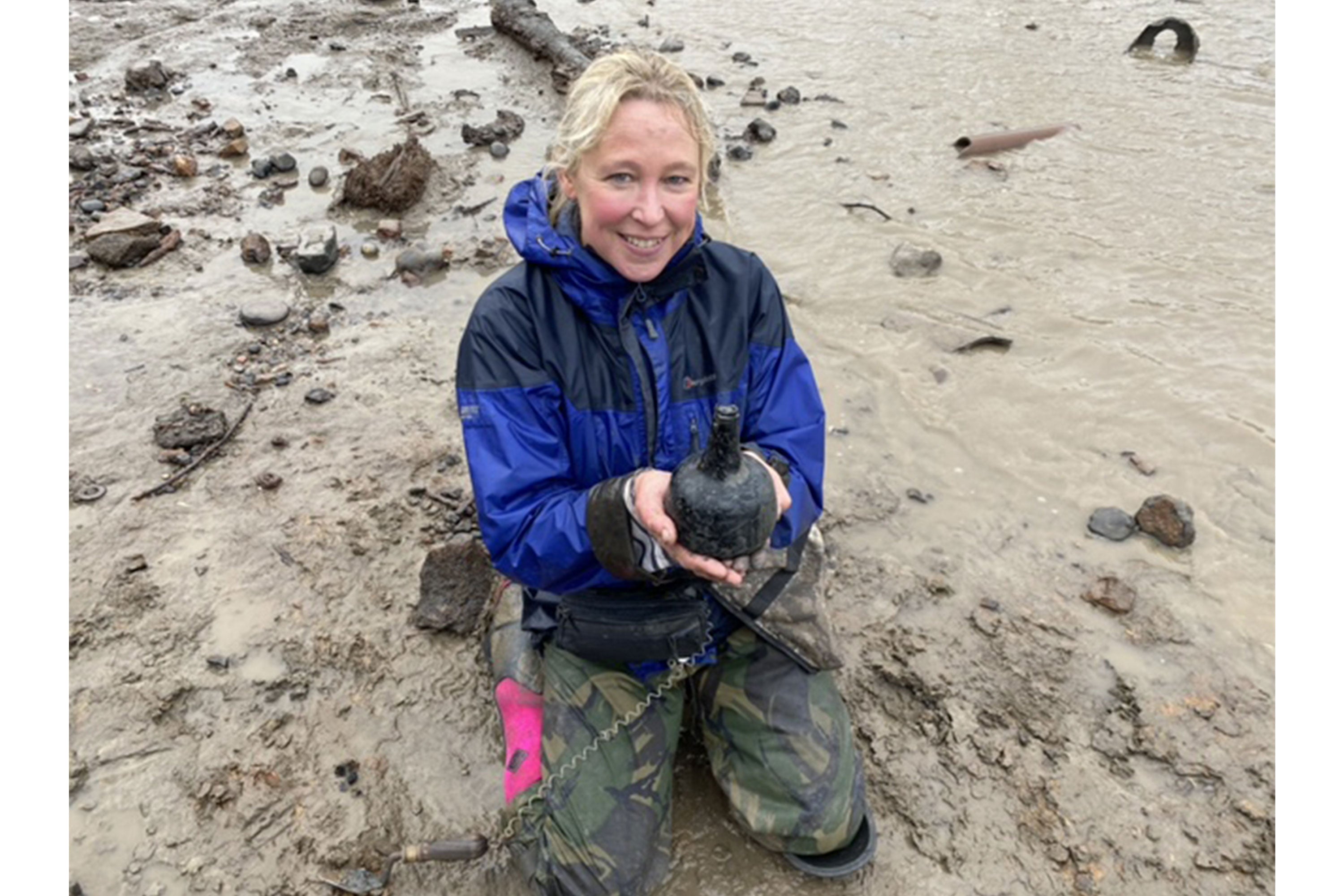 Nicola White holding the 18th Century bottle whilst crouched down on the muddy shoreline, wearing a pair of Muck Boots