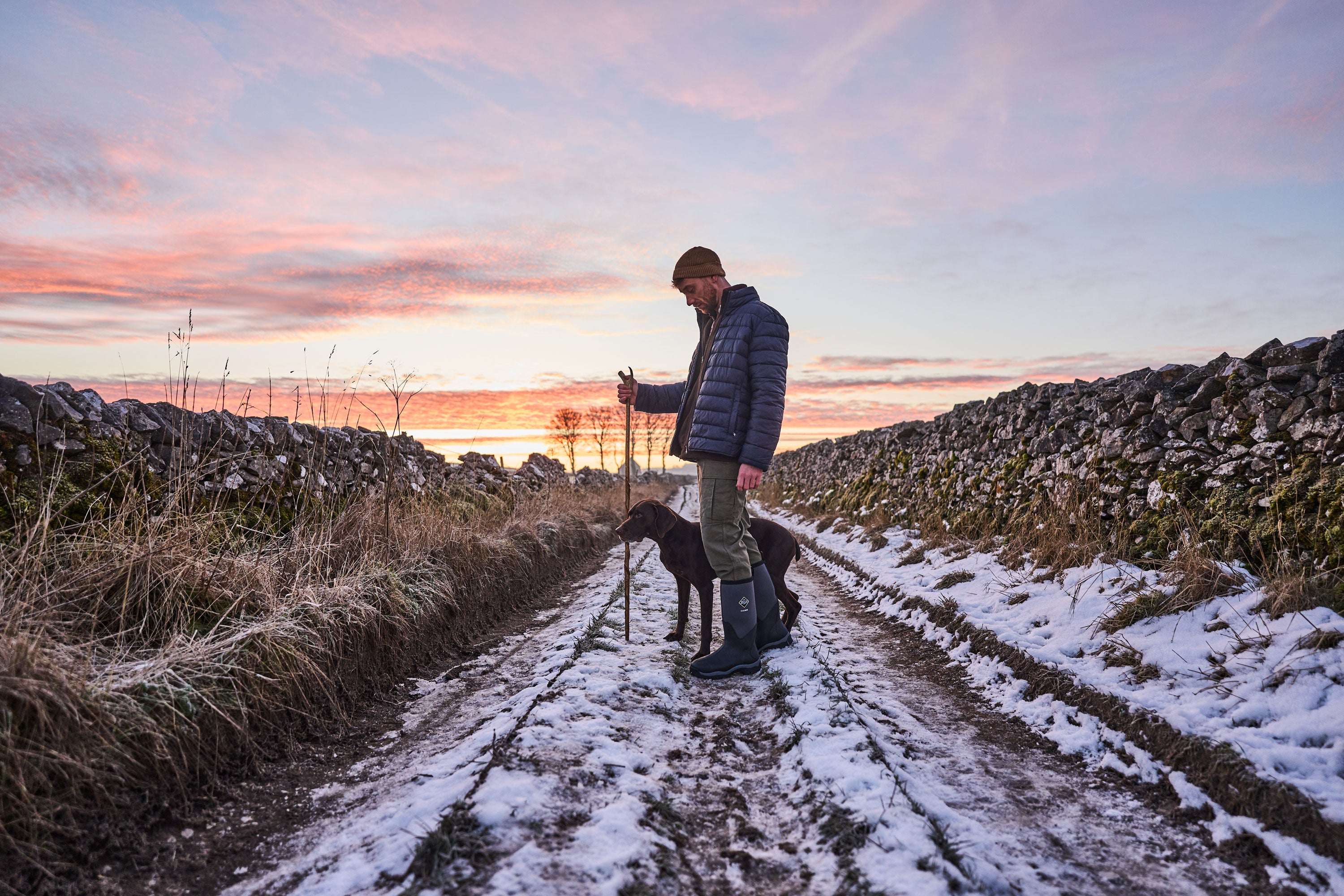 Man stood with his dog on a snow covered country lane, wearing a pair of Muck Boots Calder wellingtons