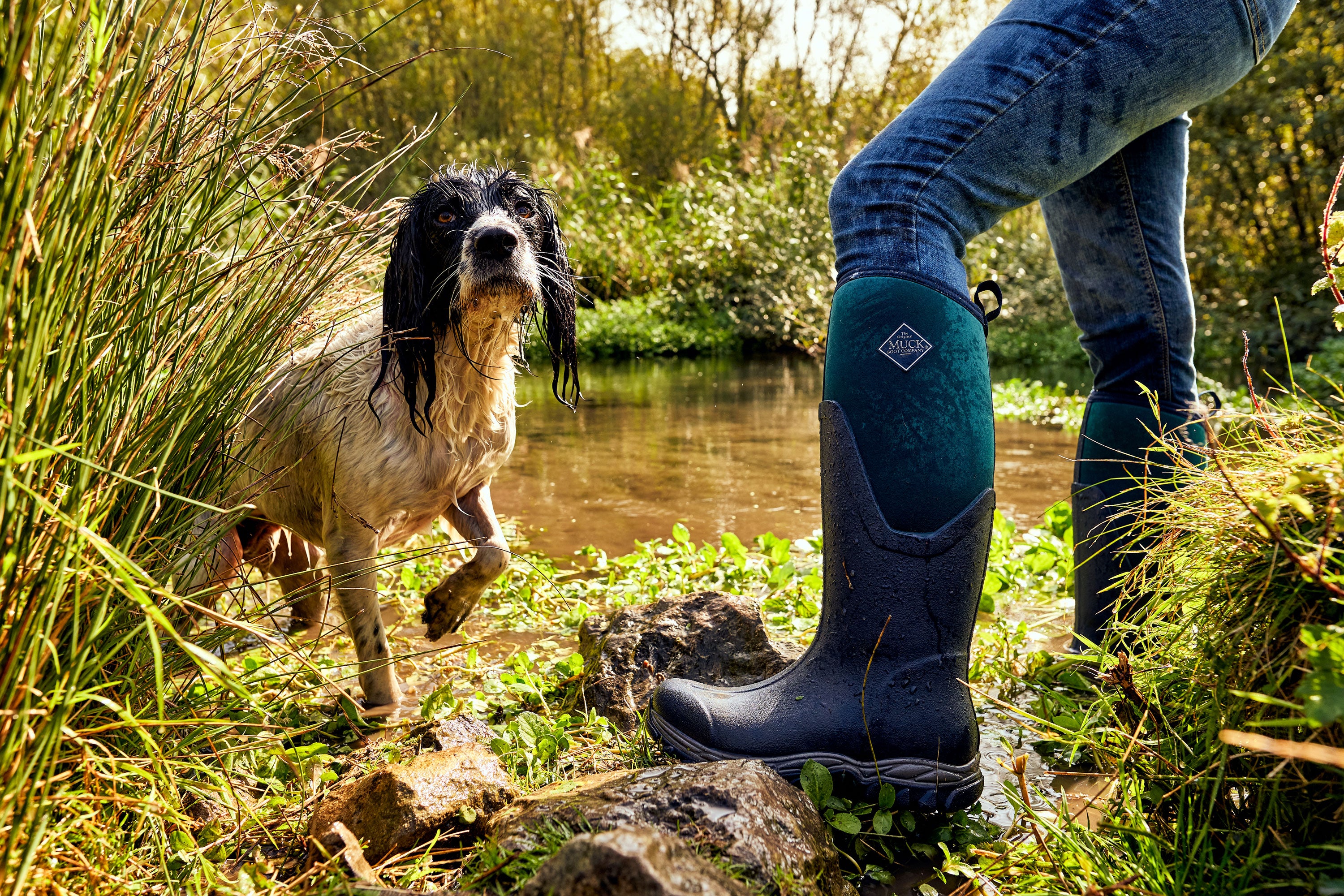 Person wearing a pair of Muck Boots Arctic Sport II Wellingtons, stood next to a stream with a wet dog