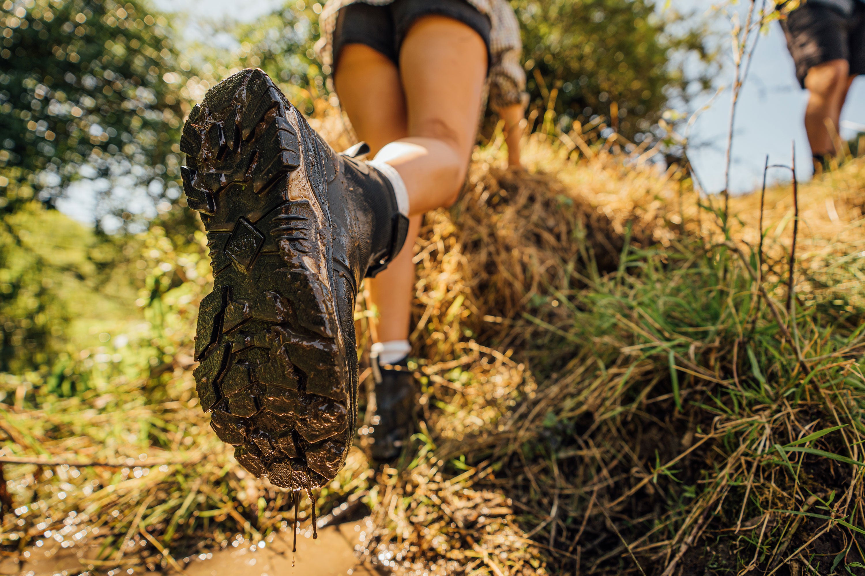 View from behind of a woman climbing through muddy ground and hay, showing the outsole of her Muck Boot Apex boots