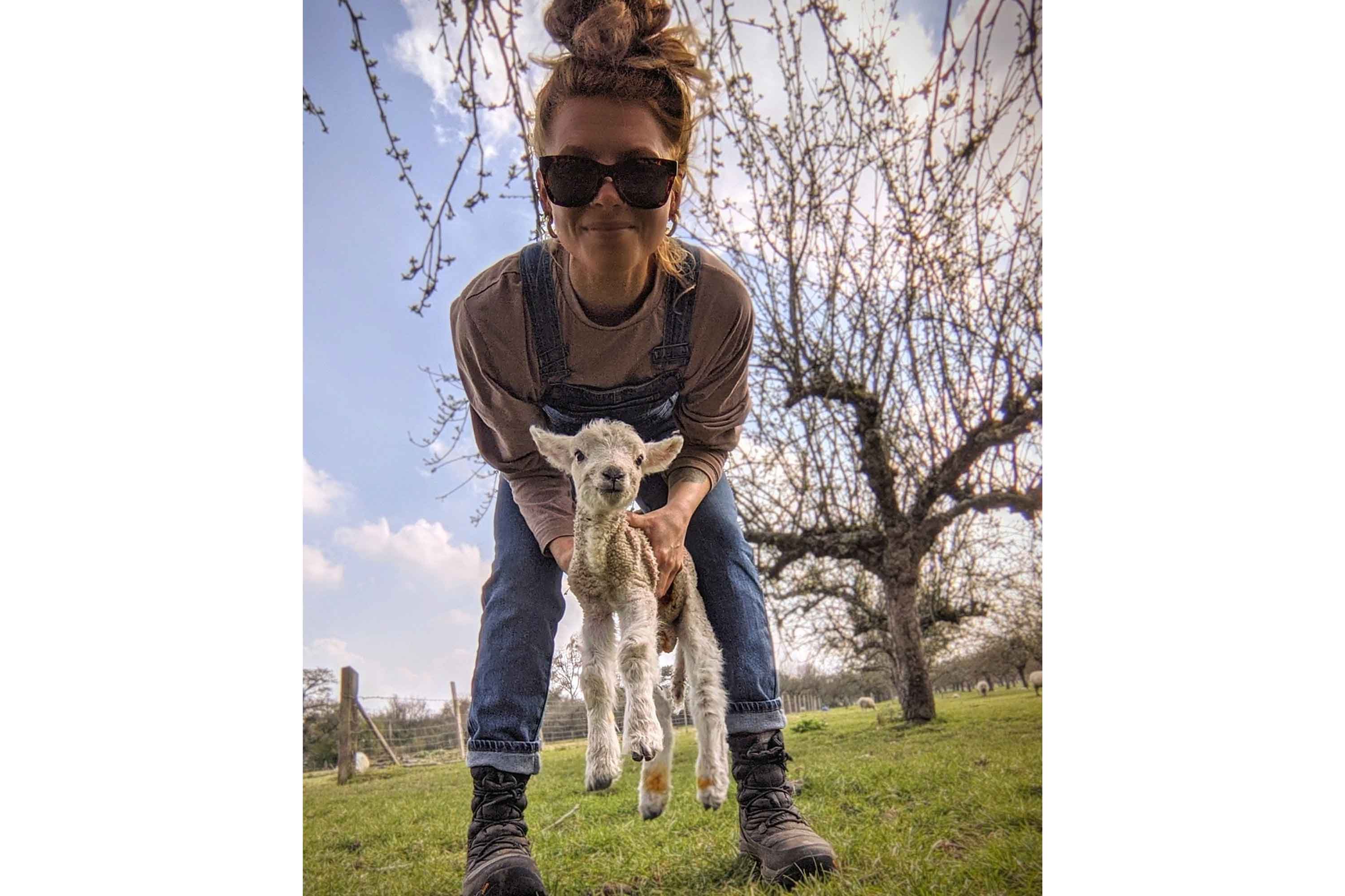 Zoe Colville stood outside in front of a blossoming tree, wearing sunglasses, dungarees and a pair of Muck Boots, holding a newly born lamb