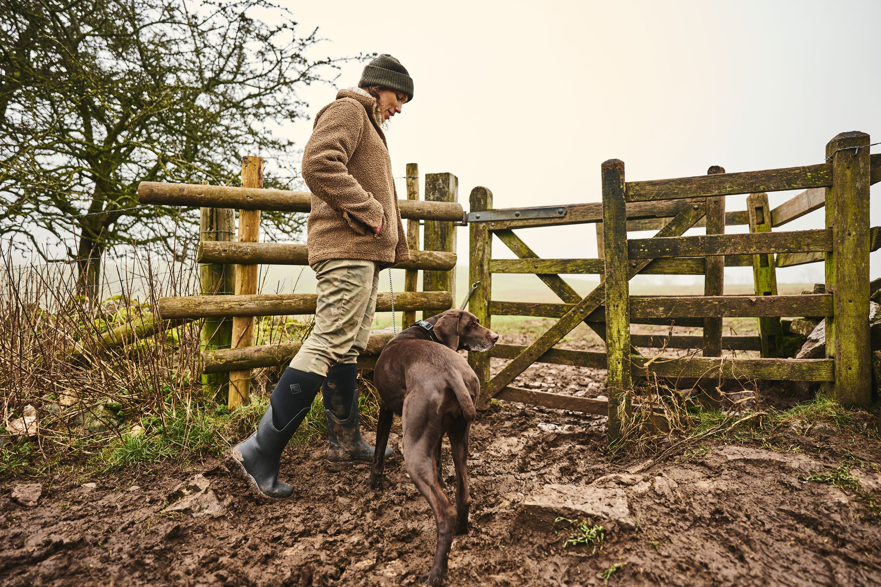 Eine Frau, die eine Mütze, einen Wollfleece, eine beige Hose und ein Paar Muck Boots trägt, führt einen braunen Hund durch ein schlammiges Feld zu einem Holztor.