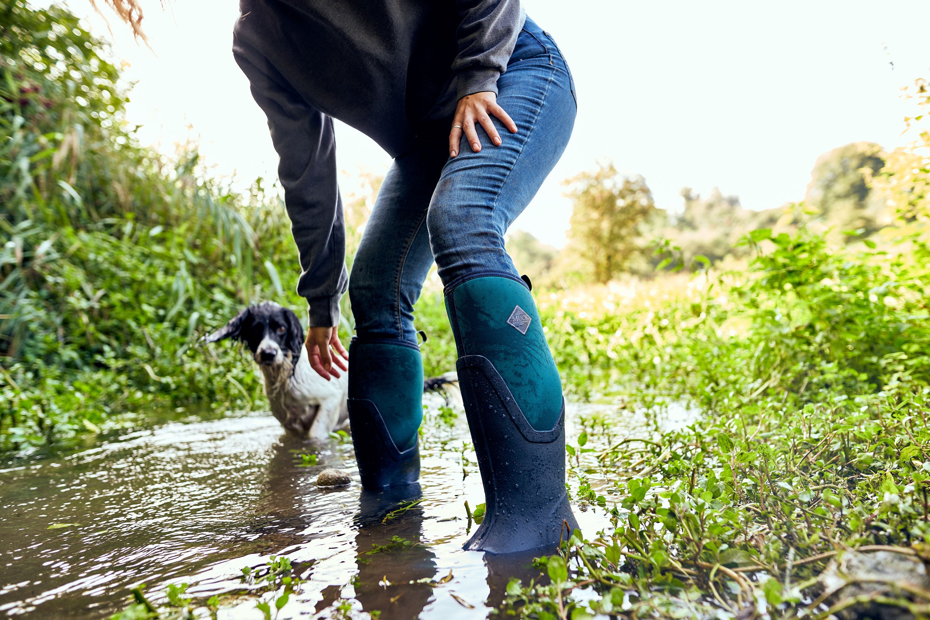 Eine nach vorn gebeugte Frau in einem Paar Muck Boots auf einem überschwemmten Weg mit Vegetation um sie herum und einem schwarz-weißen Hund im Hintergrund