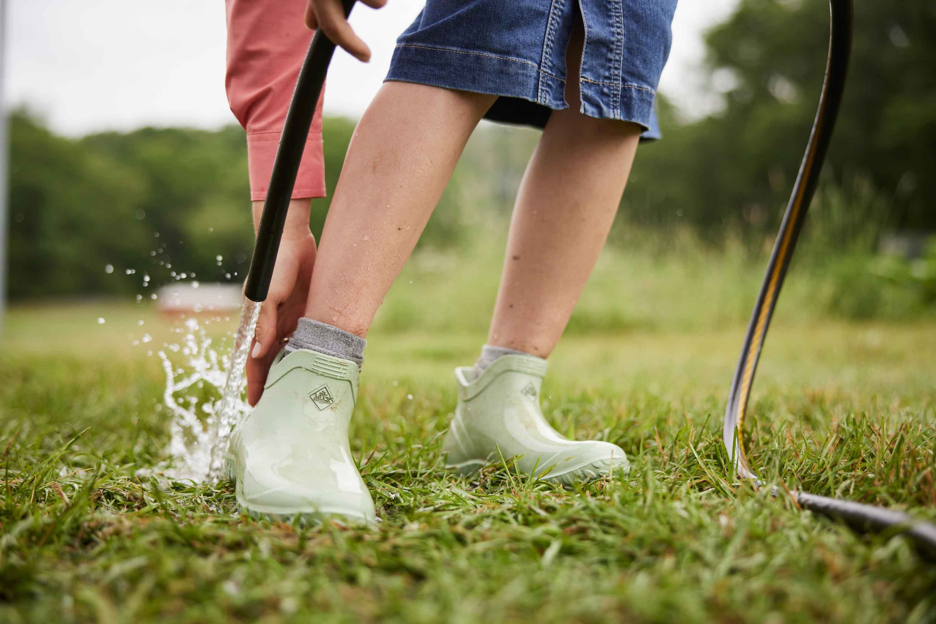 Close up of a person wearing a pair of green Muck Boot Muckster Lite Boots, watering the bottom of their heel with a hosepipe and holding the top of the heel