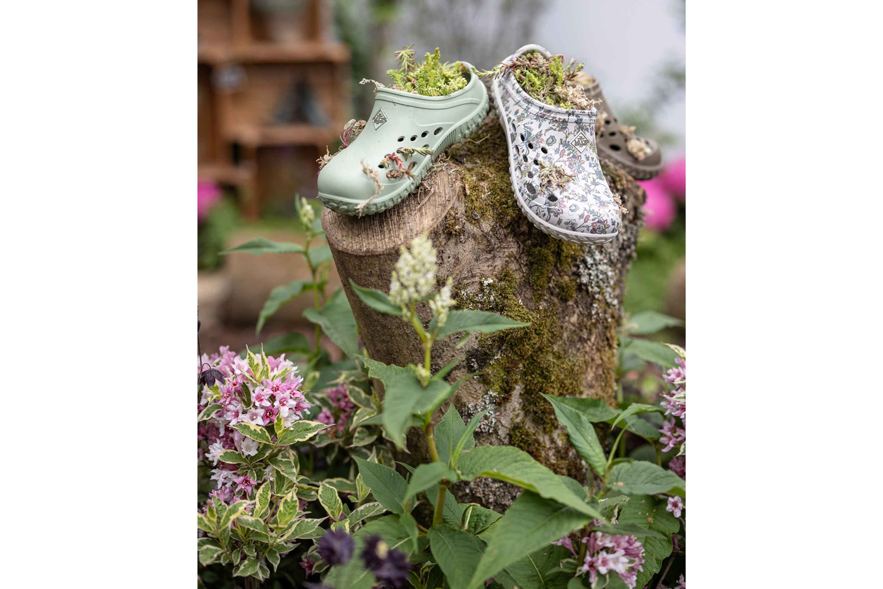 Mit Moos gefüllte Muck Boots Muckster Clogs, die auf einem von Blumen umgebenen Baumstamm ausgestellt sind