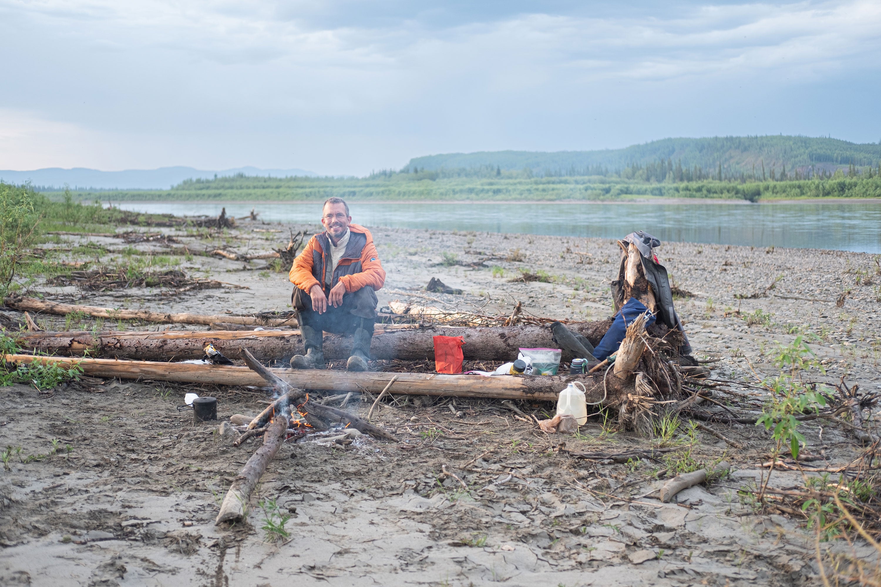 Christian sat on a log with a fire burning in front and a river with forest behind