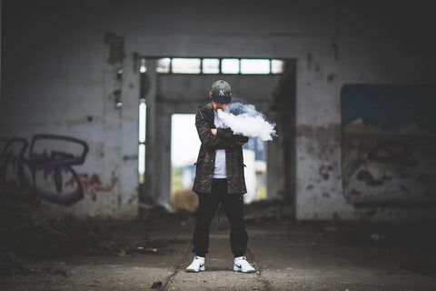 A man smoking his vape product in an isolated area with graffitis on the wall.