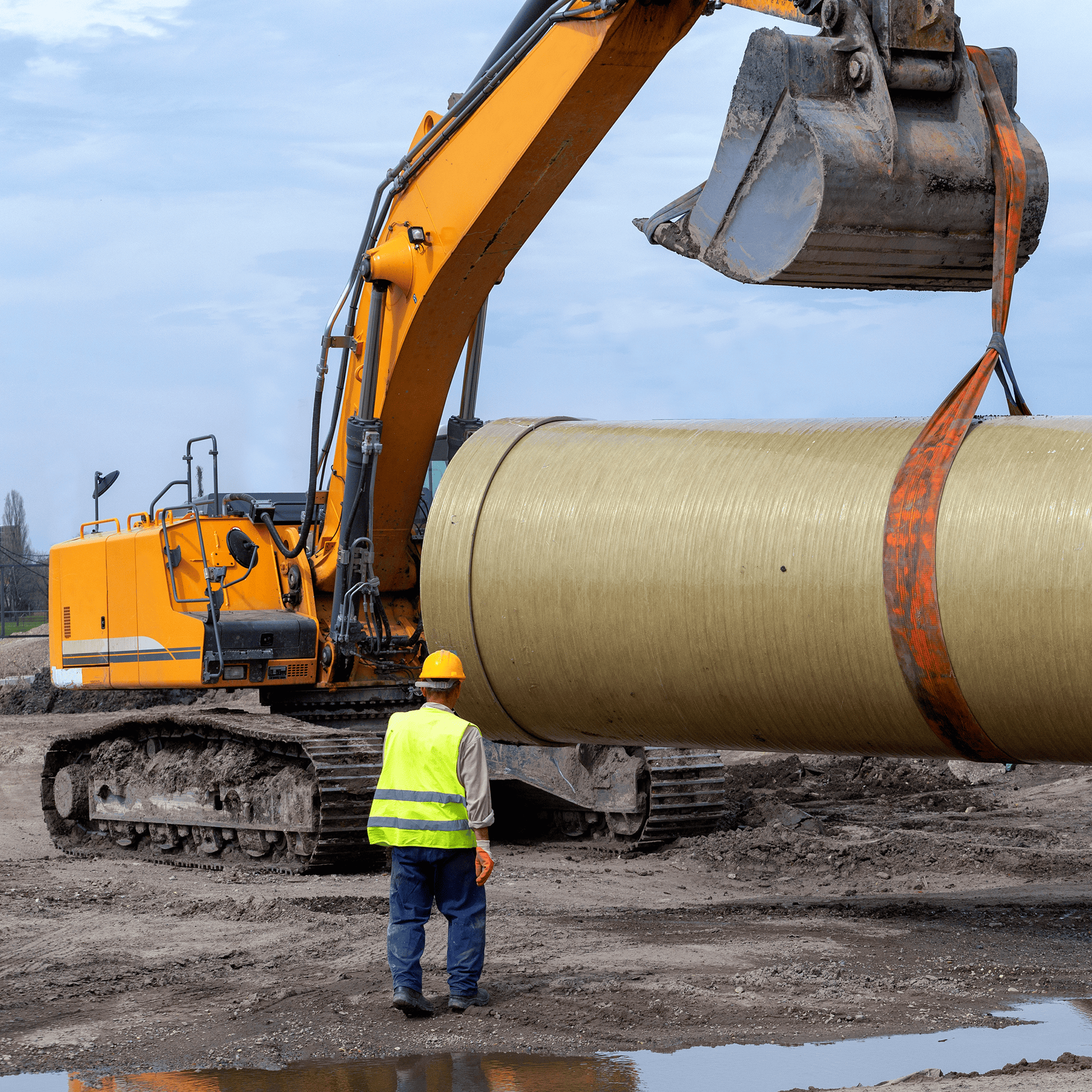 A man on a muddy jobsite walking up to an exacavator lifting a giant concrete tube via a belt secured around the arm