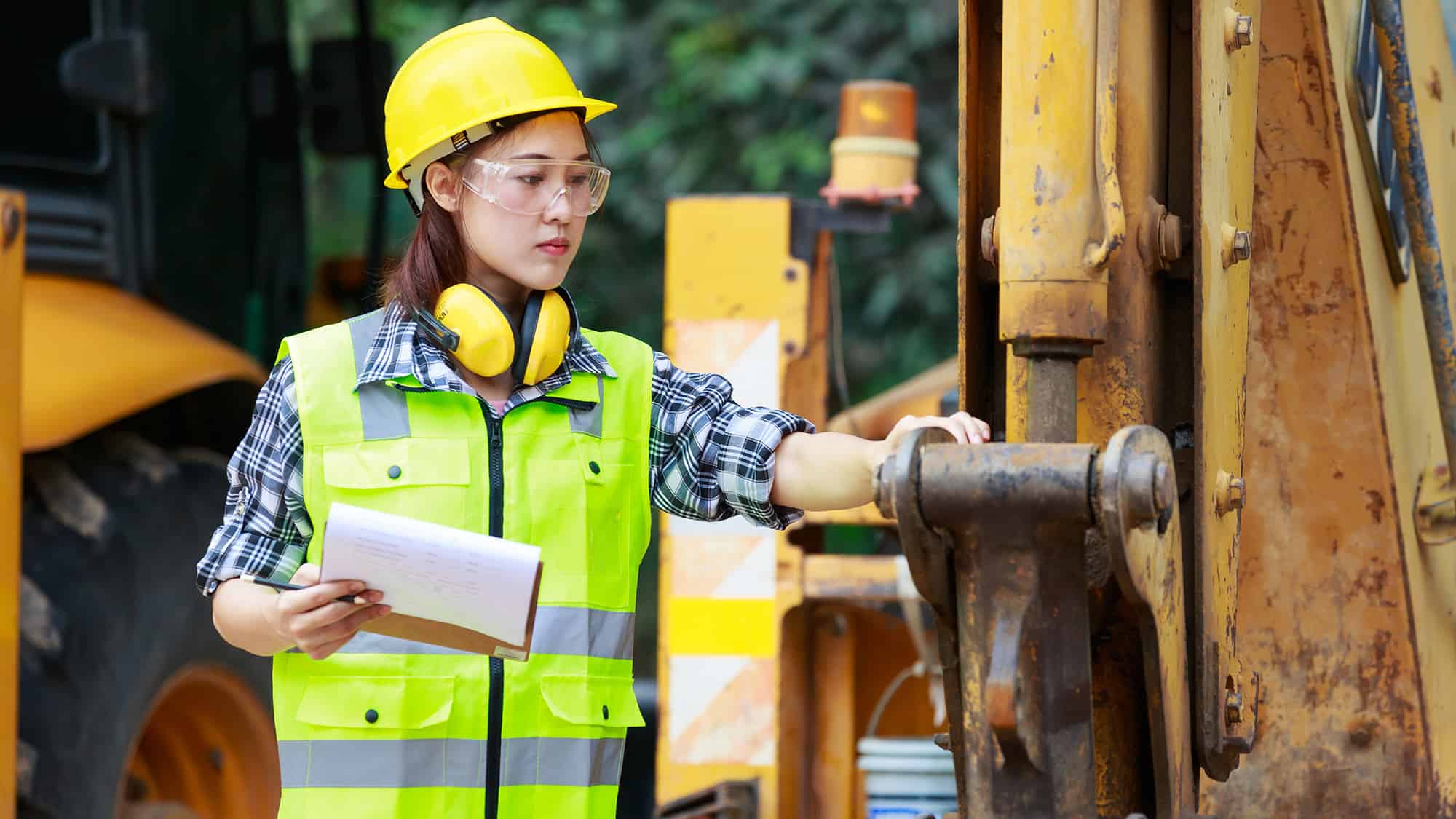 A woman wearing a safety vest and hard hat inspecting a piece of heavy equipment