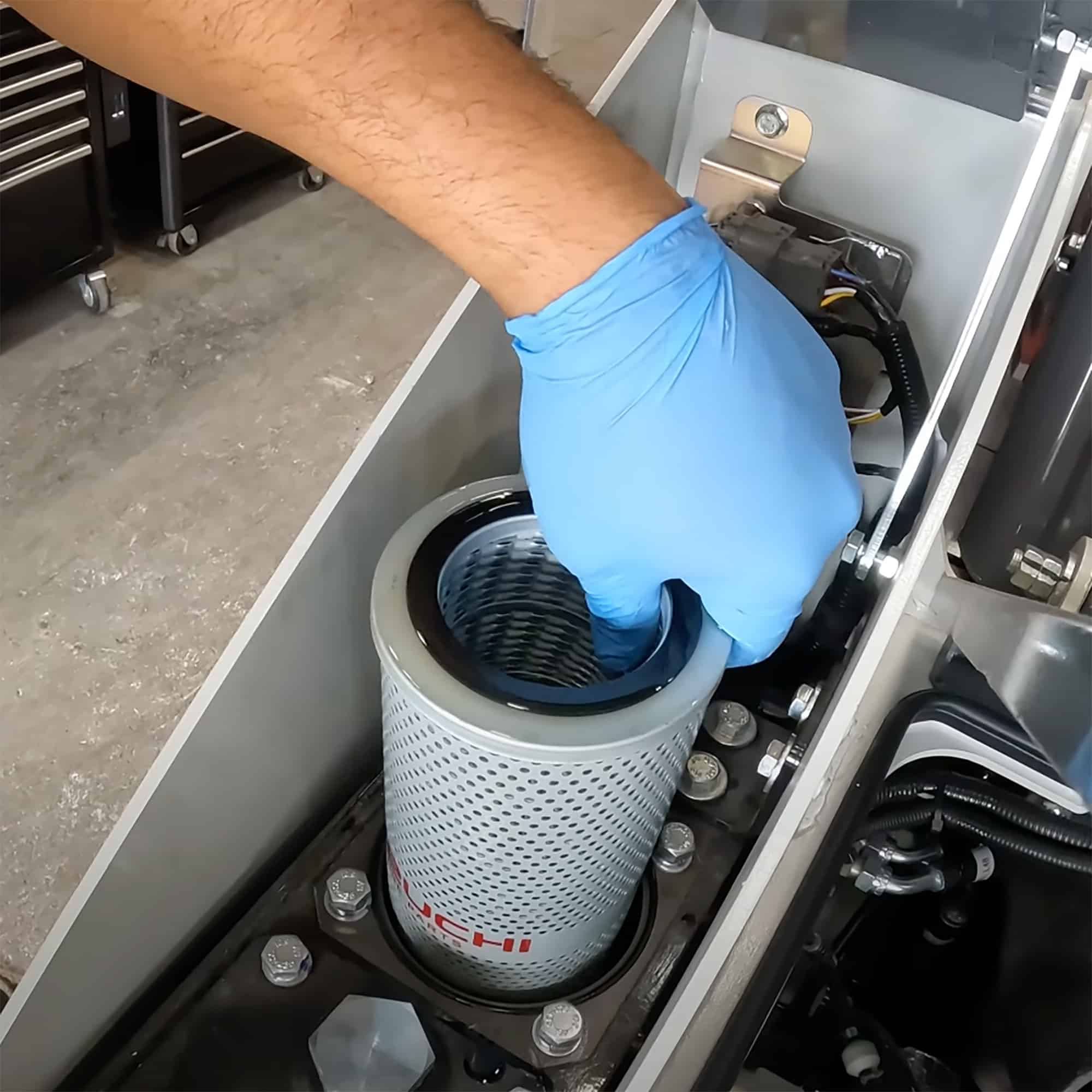 A hand wearing a blue glove reaching into a machine to check a hydraulic filter