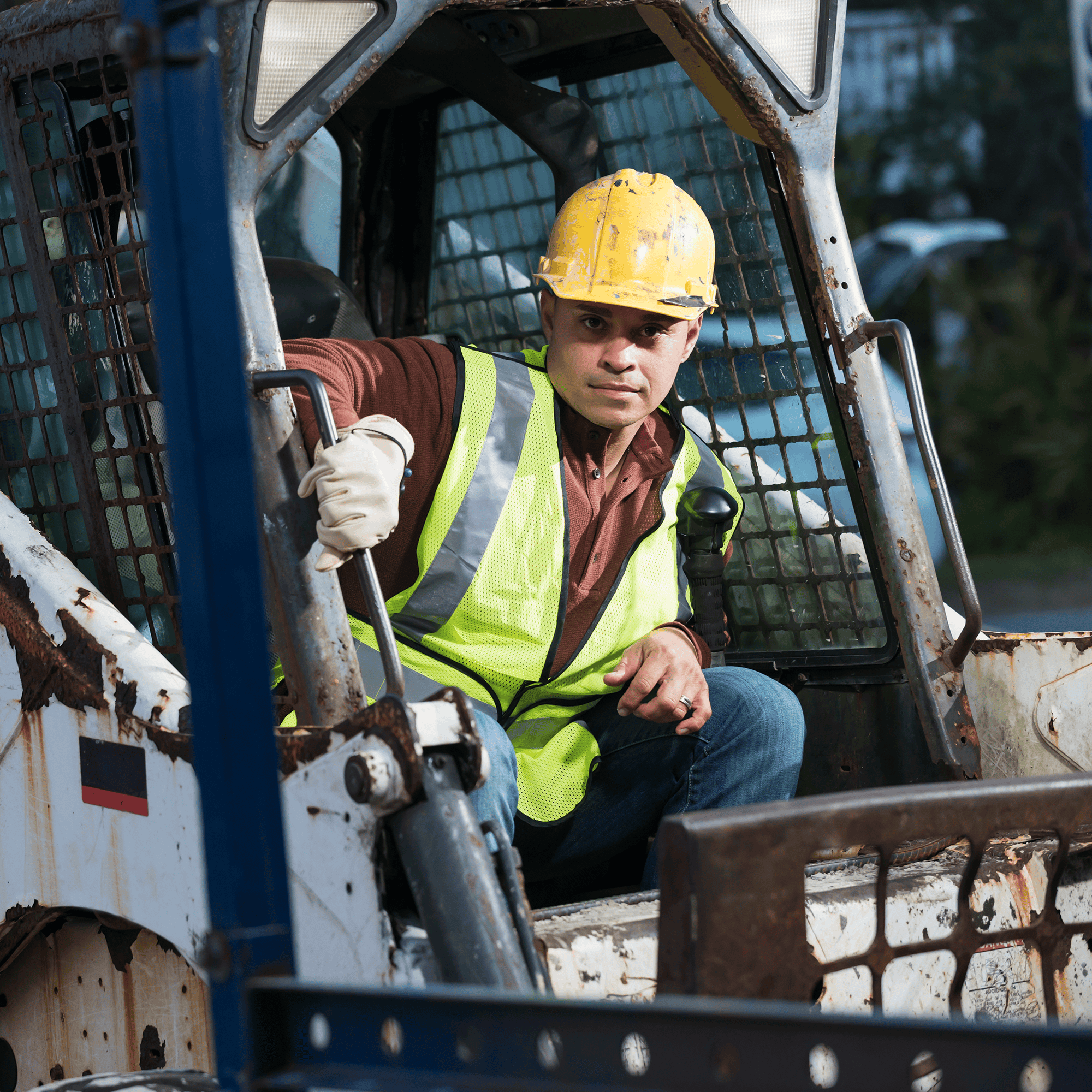 Man wearing safety vest and hard hat sitting in the cap of a skid steer
