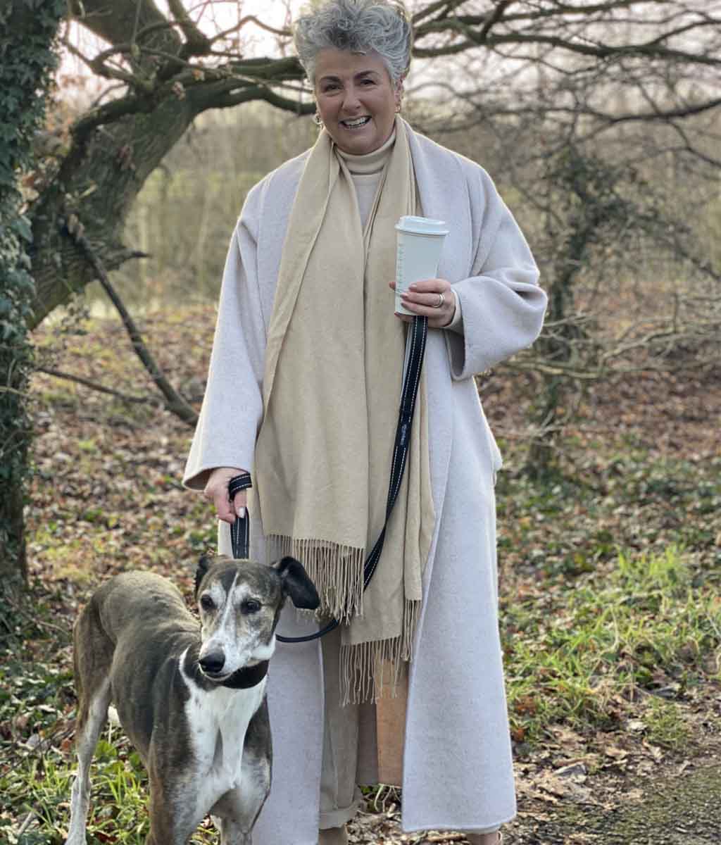 Photo showing Maxine Laceby standing outside by a tree, she is smiling and wearing a long cream coat and holding a reusable coffee cup, and her lurcher Rosie is standing by her