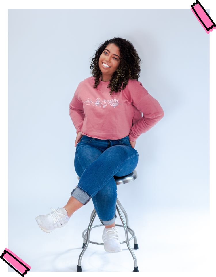 Sam smiling, sitting on a stool wearing a crewneck and jeans