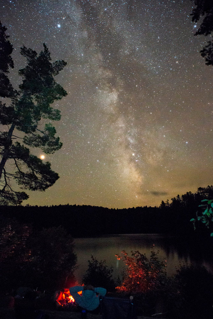 a shot of the milky way above a lake and a campfire