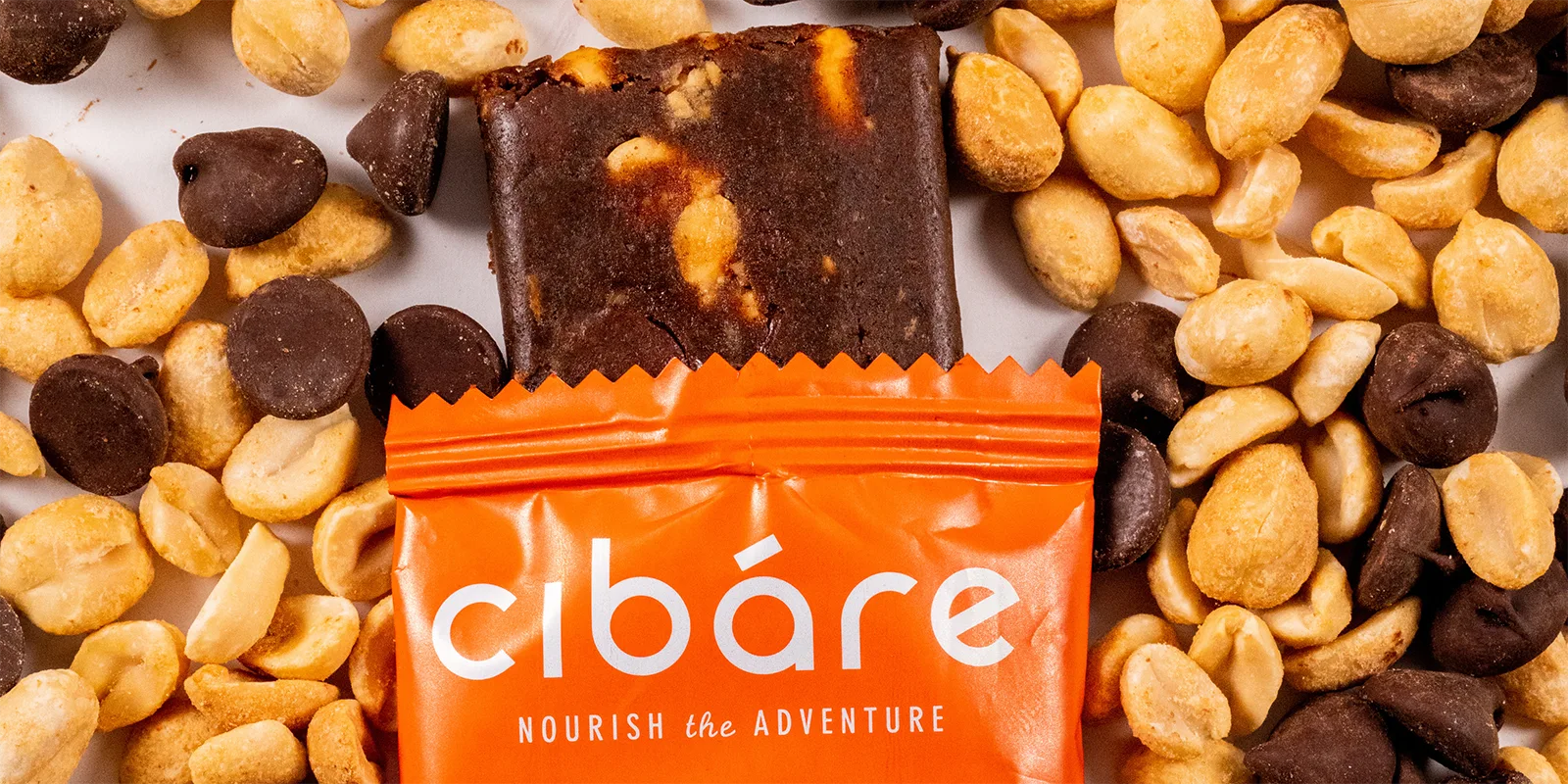 A chocolate protein bar partially wrapped in bright orange packaging with text, surrounded by roasted peanuts and chocolate chips on a white surface.