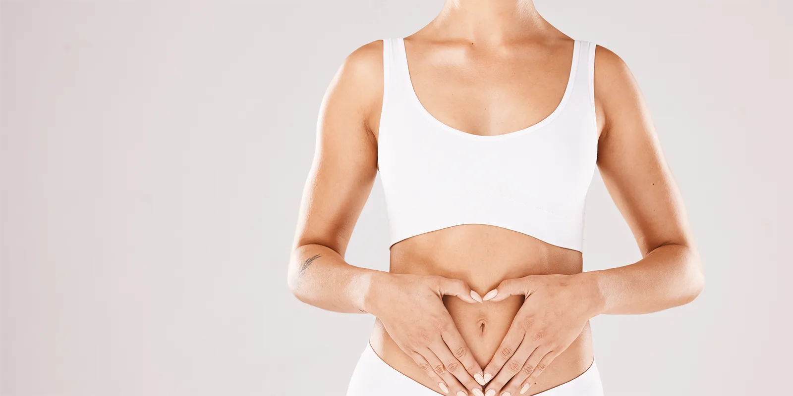 A person in a white sports bra forms a heart shape with hands over their stomach, symbolizing wellness and self-care against a neutral background.