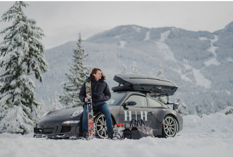 Porsche 911 with ski box in the snow with a man leaning on it with skis 