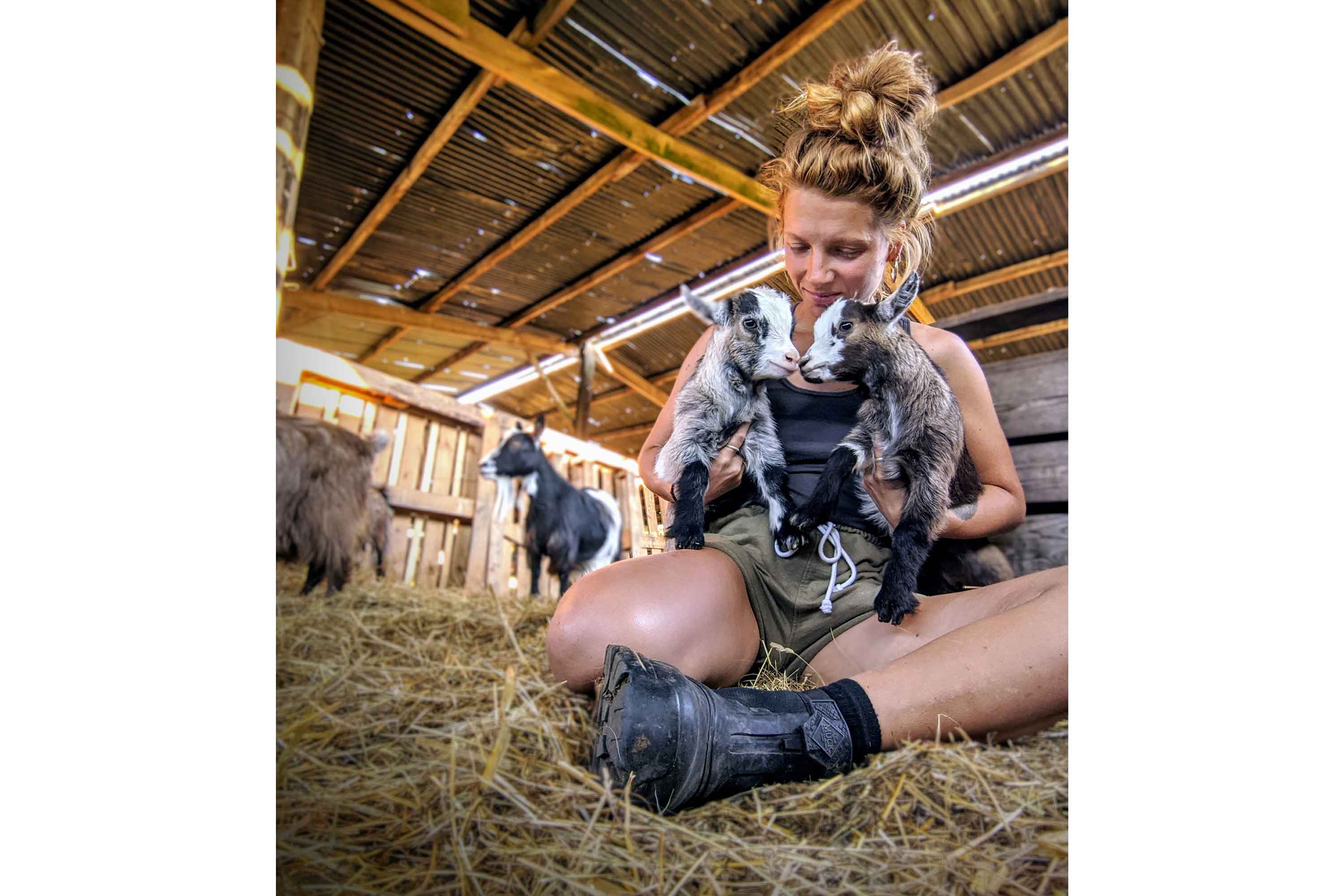Zoë Colville sat in a barn, wearing a t-shirt, shorts and pair of Muck Boot short boots, surrounded by goats and holding two goat kids