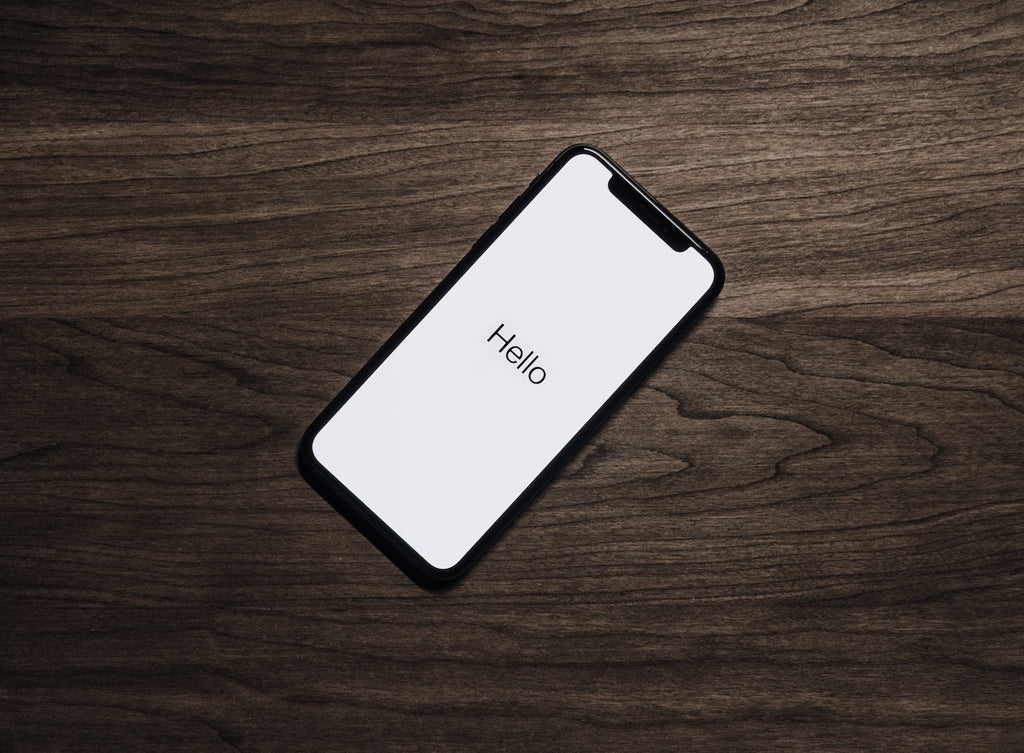 iPhone on dark wooden table surface with the welcome screen lit up displaying the word hello